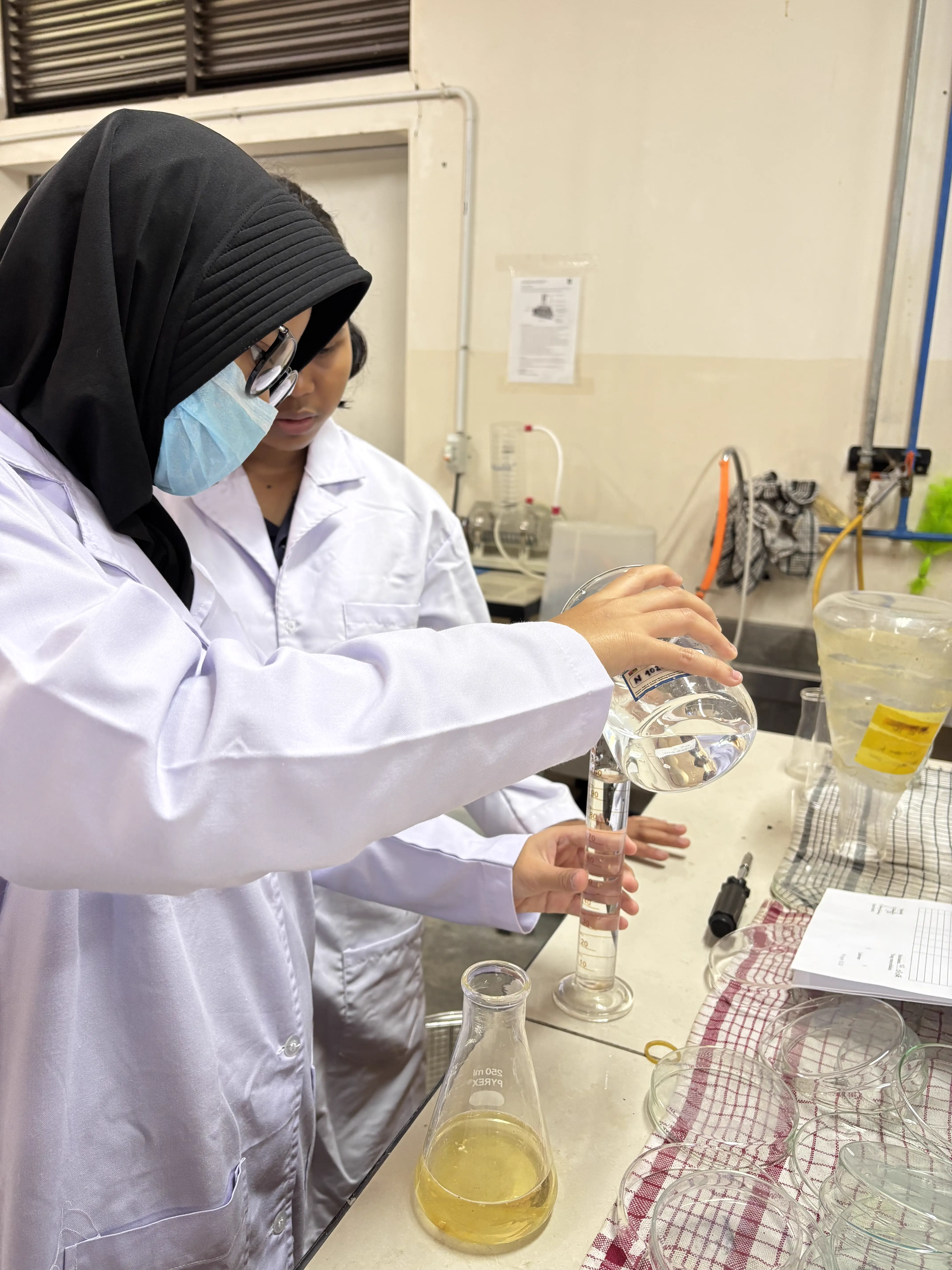 Dua siswi peneliti berhijab dan berjas lab menuangkan reagen ke dalam gelas ukur